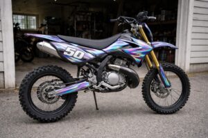 Moto 50cc de type cross garée devant un garage ouvert. La scène est prise de côté, à hauteur d’homme, avec un cadrage simple et légèrement imparfait, comme une photo prise à la main.