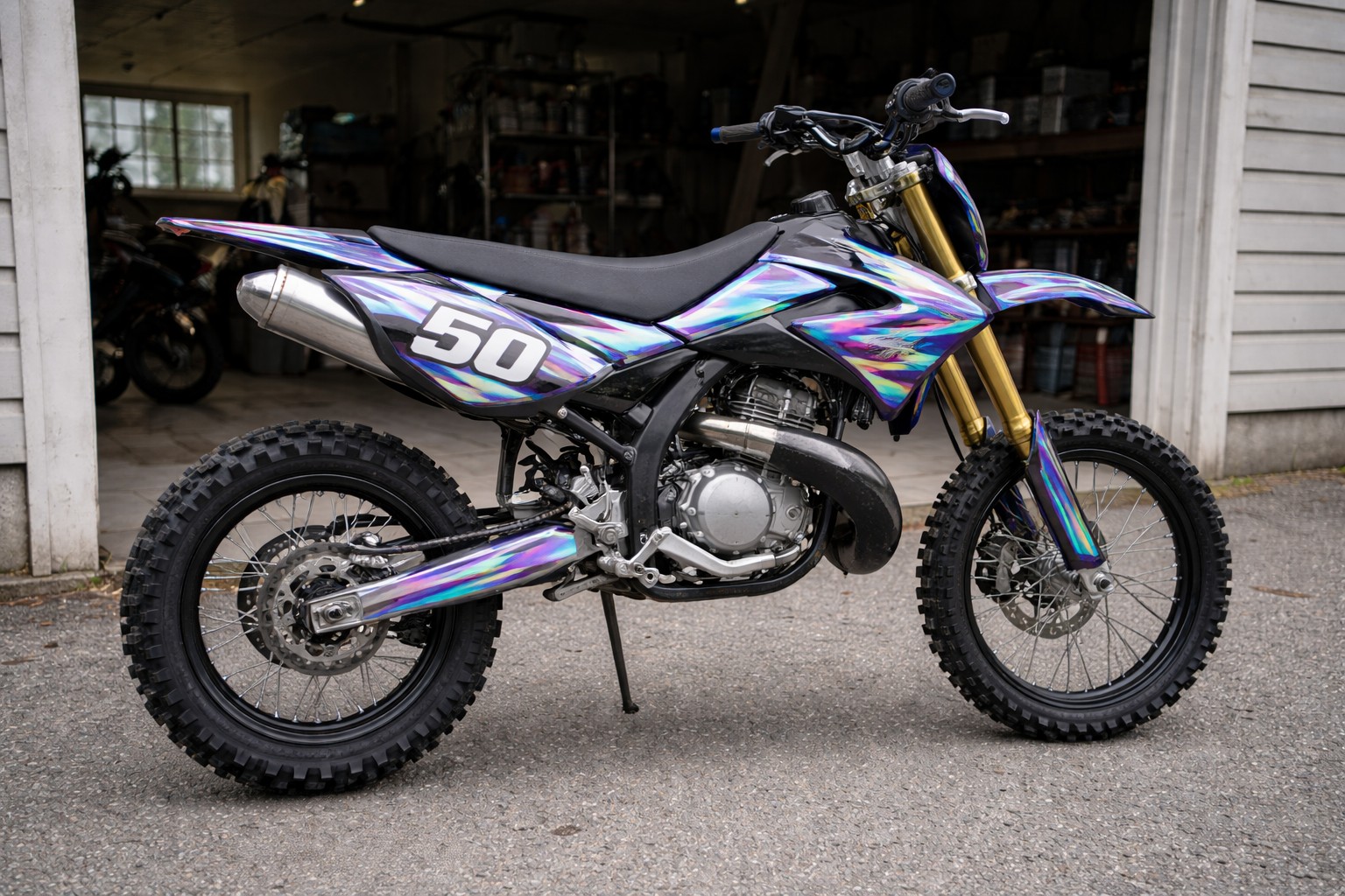 Moto 50cc de type cross garée devant un garage ouvert. La scène est prise de côté, à hauteur d’homme, avec un cadrage simple et légèrement imparfait, comme une photo prise à la main.