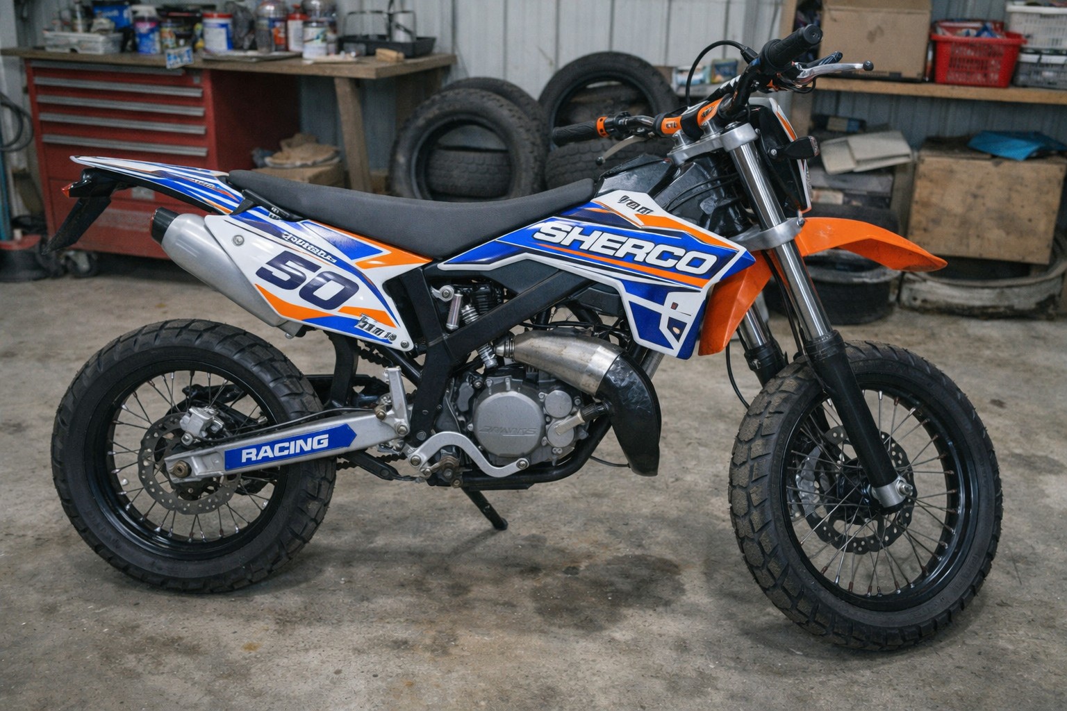 Photo d’une Sherco 50cc équipée d’un kit déco personnalisé bleu, orange et blanc, prise en vue latérale dans un garage, avec carénage custom, jantes noires, selle noire et échappement apparent, style photo réaliste Panasonic GH4