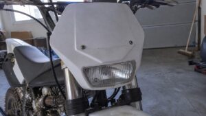 Plaque phare avant blanche pour moto 50cc type Derbi Senda enduro, avec phare rectangulaire intégré, carénage plastique légèrement sale, fourche inversée et guidon visibles en atelier.