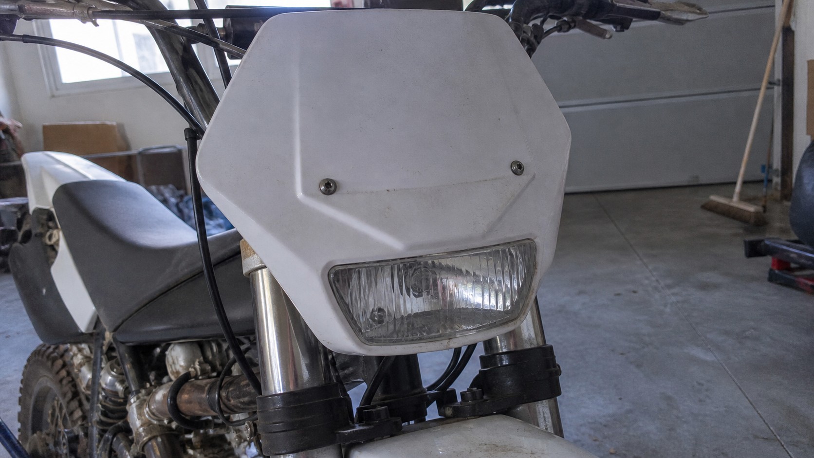 Plaque phare avant blanche pour moto 50cc type Derbi Senda enduro, avec phare rectangulaire intégré, carénage plastique légèrement sale, fourche inversée et guidon visibles en atelier.