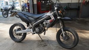 Photo réaliste d’une Derbi 50cc d’occasion de type supermotard noir et gris stationnée dans un atelier moto, avec pneus route, jantes à rayons, frein à disque, moteur apparent et autres deux-roues visibles en arrière-plan pour comparer les modèles et les prix sur le marché de l’occasion.