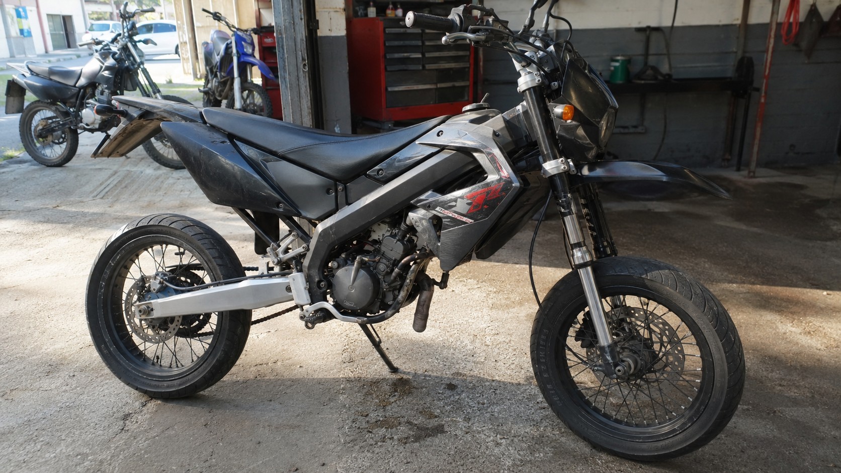 Photo réaliste d’une Derbi 50cc d’occasion de type supermotard noir et gris stationnée dans un atelier moto, avec pneus route, jantes à rayons, frein à disque, moteur apparent et autres deux-roues visibles en arrière-plan pour comparer les modèles et les prix sur le marché de l’occasion.