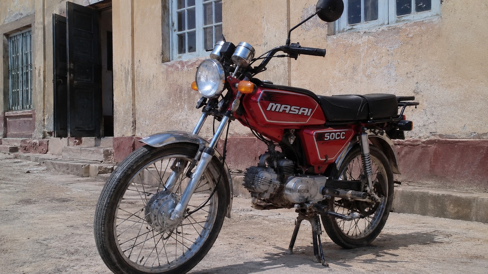 Cette image montre une Masai 50cc rouge au design rétro photographiée dans une rue ancienne sous une lumière naturelle ensoleillée. On distingue clairement le réservoir rouge siglé MASAI, le moteur monocylindre 50 cm³, les jantes à rayons, les suspensions arrière chromées et la selle noire biplace. L'arrière-plan composé d'un bâtiment ancien aux façades usées apporte un contexte urbain authentique. Cette photographie illustre parfaitement une moto 50cc de type utilitaire vintage destinée aux déplacements quotidiens en ville.