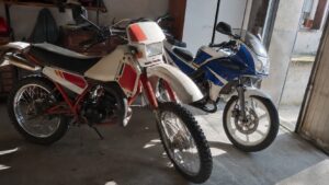 Photo réaliste de deux motos 50cc Yamaha style DT et TZR dans un garage ensoleillé, avec une mécaboîte japonaise visible, lumière naturelle, sol béton et ambiance atelier mécanique authentique