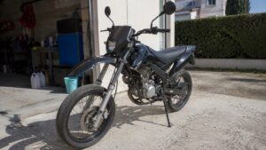Moto 50cc mécaboîte noire garée sur une allée bétonnée devant un garage domestique, photographiée en lumière naturelle avec détails visibles du moteur, du guidon, des rétroviseurs, de la fourche, des roues à rayons et du sélecteur de vitesse, pour un article expliquant pourquoi choisir une mécaboîte pour débuter.