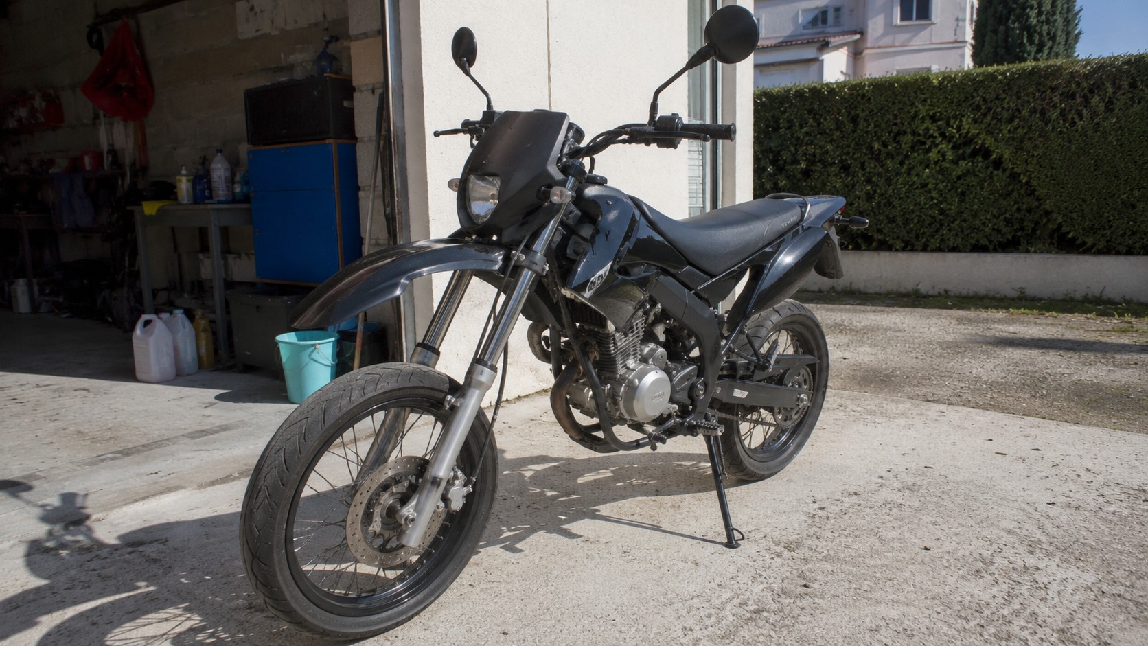 Moto 50cc mécaboîte noire garée sur une allée bétonnée devant un garage domestique, photographiée en lumière naturelle avec détails visibles du moteur, du guidon, des rétroviseurs, de la fourche, des roues à rayons et du sélecteur de vitesse, pour un article expliquant pourquoi choisir une mécaboîte pour débuter.