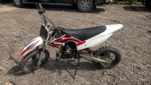 Moto cross 50cc d'occasion blanche et rouge photographiée en extérieur sur un terrain gravillonné sous un temps ensoleillé, vue latérale gauche montrant les pneus tout-terrain à crampons, le moteur monocylindre, la transmission par chaîne, la suspension arrière rouge et les carénages sportifs. Photo réaliste type annonce de vente de moto cross 50cc d'occasion.