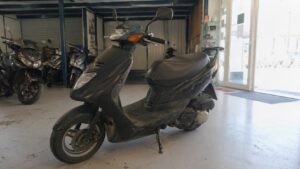Scooter 50cc d’occasion noir, modèle urbain compact avec carénage avant arrondi, clignotants orange, selle biplace, rétroviseurs et traces d’usure, photographié dans un garage showroom de scooters.