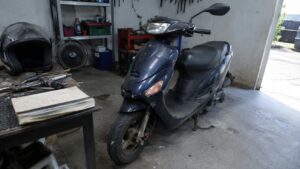 Photo réaliste d’un scooter 50cc d’occasion dans un atelier de réparation encombré, avec carrosserie rayée, pneus usés, outils, casque et documents d’entretien, illustrant les risques d’une fausse bonne affaire lors de l’achat d’un scooter pas cher.