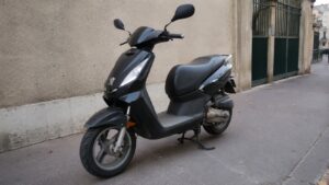 Scooter Peugeot Ludix 50cc noir stationné sur un trottoir en ville, avec logo Peugeot visible, selle noire, rétroviseurs, roue avant et carrosserie scooter 50cc d’occasion