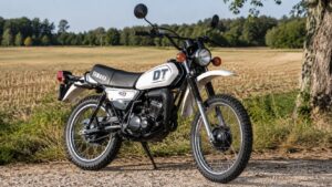Photo professionnelle ultra réaliste au format 16/9 d’une Yamaha DT 50cc blanche vintage, moto enduro 50cc garée sur un chemin de campagne ensoleillé, avec champ doré, arbre et arrière-plan naturel légèrement flou.