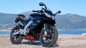 Photo professionnelle au format 16/9 d’une Aprilia RS 50cc noire garée en extérieur par temps ensoleillé, avec vue sur la mer, les montagnes en arrière-plan et une lumière naturelle mettant en valeur les détails de la moto sportive.