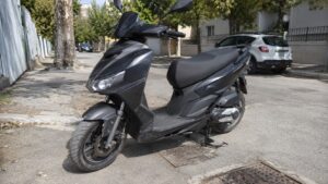 Aprilia SXR 50 noir stationné sur une rue urbaine ensoleillée, scooter 50cc vu de trois quarts avec carrosserie anguleuse, selle longue noire, roue avant visible, béquille centrale, chaussée avec plaques d’égout, arbres, voitures garées et immeubles résidentiels en arrière-plan, photo amateur style Panasonic GH4, 10 juillet 2026, format 16/9.