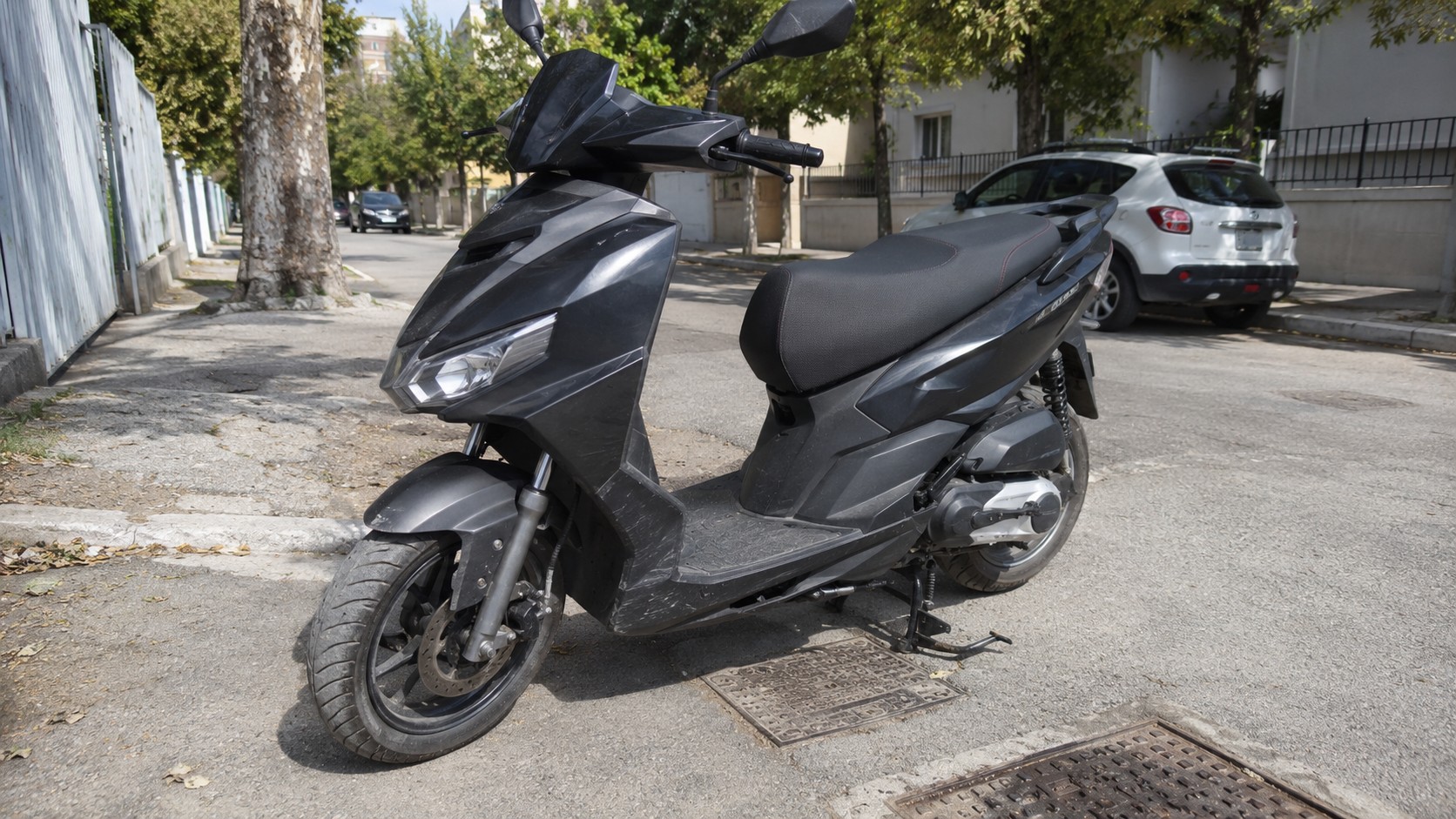 Aprilia SXR 50 noir stationné sur une rue urbaine ensoleillée, scooter 50cc vu de trois quarts avec carrosserie anguleuse, selle longue noire, roue avant visible, béquille centrale, chaussée avec plaques d’égout, arbres, voitures garées et immeubles résidentiels en arrière-plan, photo amateur style Panasonic GH4, 10 juillet 2026, format 16/9.