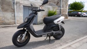 MBK Stunt 50cc noir et blanc stationné sur une rue urbaine devant une façade en pierre, scooter équipé de jantes noires, frein à disque avant, selle noire, rétroviseurs et carénage sportif, lumière naturelle ensoleillée, juillet 2026, format 16:9