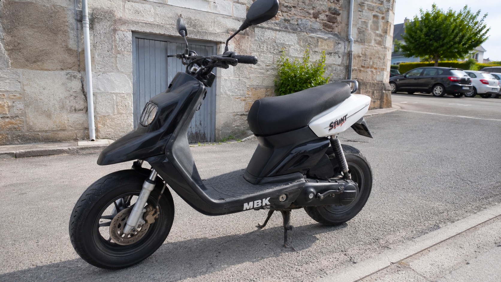 MBK Stunt 50cc noir et blanc stationné sur une rue urbaine devant une façade en pierre, scooter équipé de jantes noires, frein à disque avant, selle noire, rétroviseurs et carénage sportif, lumière naturelle ensoleillée, juillet 2026, format 16:9