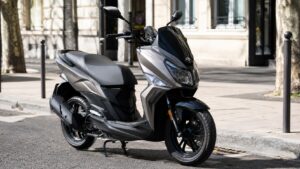 SYM Jet 14 50cc gris et noir, scooter maxi-look 125, stationné en rue urbaine ensoleillée, façade claire, arbres et mobilier urbain, vue trois-quarts avant, 29-07-2026, format 16:9