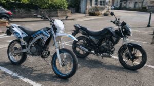deux motos 50cc stationnées sur un parking goudronné, supermotard blanc et bleu avec roues à rayons et roadster noir avec pneus routiers, comparaison de modèles adaptés au bitume, village français en arrière-plan, 22 juillet 2026, format 16:9
