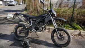 débridage_50cc_vitesse_réelle_loi_risques_2026_moto_type_supermotard_50cc_noire_moteur_apparent_casque_sur_route_urbaine_soleil_format_16_9