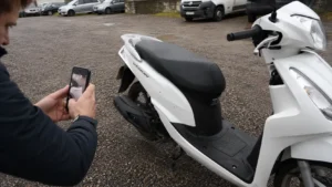 homme_photographie_smartphone_scooter_50_honda_vision_blanc_bequille_centrale_parking_gravier_humide_30_juillet_2026_panoramique_1672x941