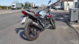 moto 50cc routière grise et rouge stationnée près d’une station-service, vue arrière trois quarts, pneu asphaltique, échappement métallique, feu arrière et moteur visibles, circulation et véhicules en arrière-plan, 22 juillet 2026, format 16:9
