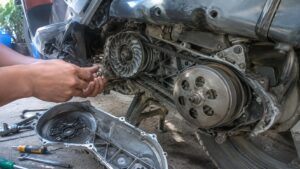 Variateur de scooter générique démonté, transmission CVT ouverte avec courroie, poulie ventilée, embrayage, carter retiré, outils au sol et mains de mécanicien pendant le réglage, 2026-07-15, format 16:9