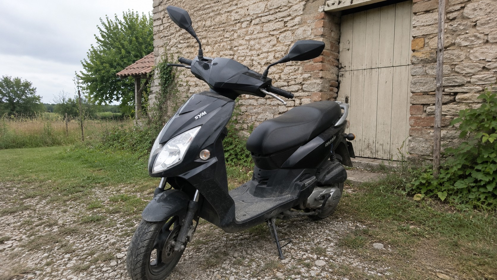 scooter_50cc_SYM_noir_garage_ancienne_grange_en_pierre_campagne_française_herbe_gravier_juillet_2026_image_16_9