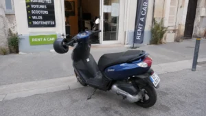 scooter_50_peugeot_kisbee_bleu_metallise_casque_jet_noir_retroviseur_devant_agence_rent_a_car_trottoir_30_juillet_2026_panoramique_1672x941