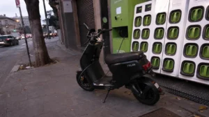 scooter_electrique_niu_noir_mat_station_echange_batteries_gogoro_verte_trottoir_dalle_ville_crepuscule_30_juillet_2026_panoramique_1672x941