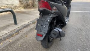 support de plaque 50cc sur scooter noir vue arrière, porte-plaque vide fixé au garde-boue, feu arrière rouge, catadioptre, roue arrière avec pneu route, amortisseur à ressort, échappement latéral, béquille, stationnement en bord de chaussée urbaine, lumière naturelle ensoleillée, photo Canon EOS R6 2026