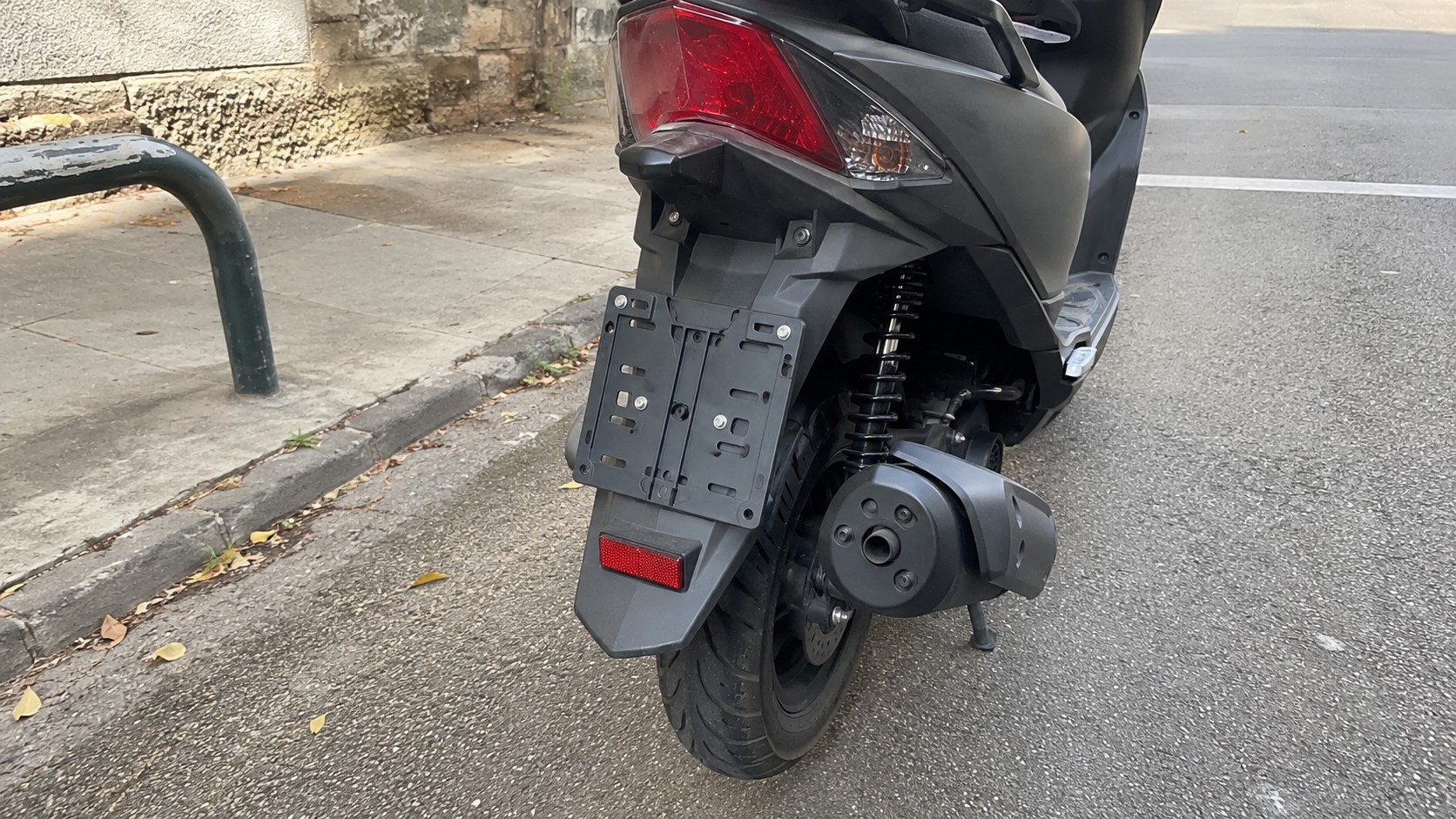 support de plaque 50cc sur scooter noir vue arrière, porte-plaque vide fixé au garde-boue, feu arrière rouge, catadioptre, roue arrière avec pneu route, amortisseur à ressort, échappement latéral, béquille, stationnement en bord de chaussée urbaine, lumière naturelle ensoleillée, photo Canon EOS R6 2026