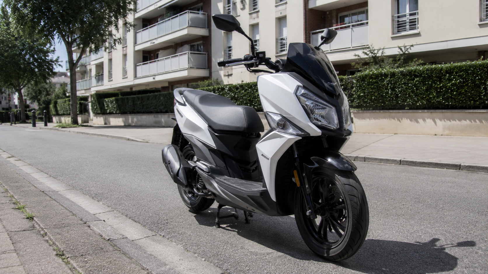 SYM Jet 14 50 cm3 blanc nacre et noir a bulle fumee vu de trois quarts avant, monocylindre 4 temps de 49 cm3 a injection electronique, stationne le long d'un trottoir