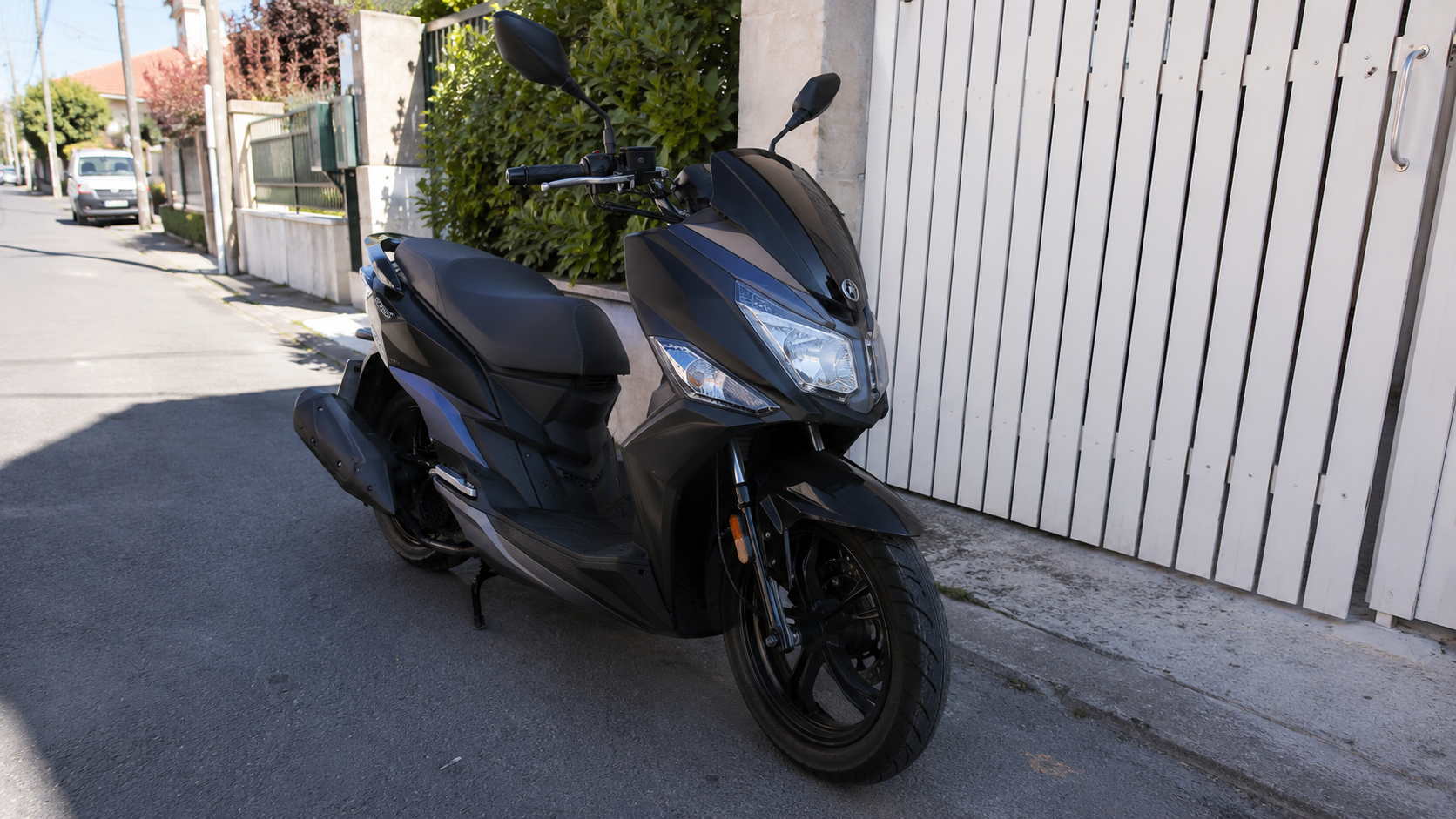 SYM Jet 14 50 cm3 gris anthracite et noir mat vu de trois quarts avant, roues de 14 pouces et disque avant de 260 mm, gare sur bequille laterale devant un portail blanc