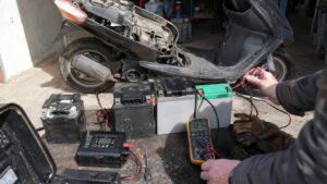 contrôle technique de plusieurs batteries de scooter 50cc plomb AGM gel et lithium devant un scooter noir démonté, mécanicien mesurant une batterie 12 V au multimètre affichant 12,74 volts, chargeur branché, ancienne batterie avec bornes sulfatées, câbles rouges et noirs, gants et outils d’entretien sur établi extérieur ensoleillé, diagnostic de panne de démarrage et préparation à l’hivernage, photographie documentaire Canon EOS R6 grand-angle 24–28 mm, lumière naturelle, août 2026