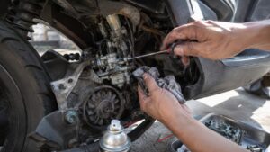 Gros plan technique d’un carburateur de scooter 50cc accessible dans le compartiment moteur ouvert, mains d’un mécanicien amateur réglant une vis du carburateur avec un tournevis rouge et noir tout en tenant un chiffon graisseux, transmission et variateur visibles à gauche, durites câbles colliers et raccords autour du carburateur, bombe nettoyante au premier plan et pièce de carburateur démontée dans un bac métallique à droite, traces de graisse et d’usure mécanique réalistes, lumière naturelle ensoleillée, arrière-plan extérieur flouté, prise de vue documentaire Canon EOS R6 grand-angle 24–28 mm, format 16:9, 2026