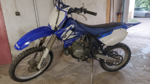 Yamaha moto cross 50cc bleue blanches vue latérale garage intérieur maison française fourche avant guidon pneus crantés jantes à rayons frein à disque moteur 2 temps chaine bras oscillant béquille lumière naturelle 2026