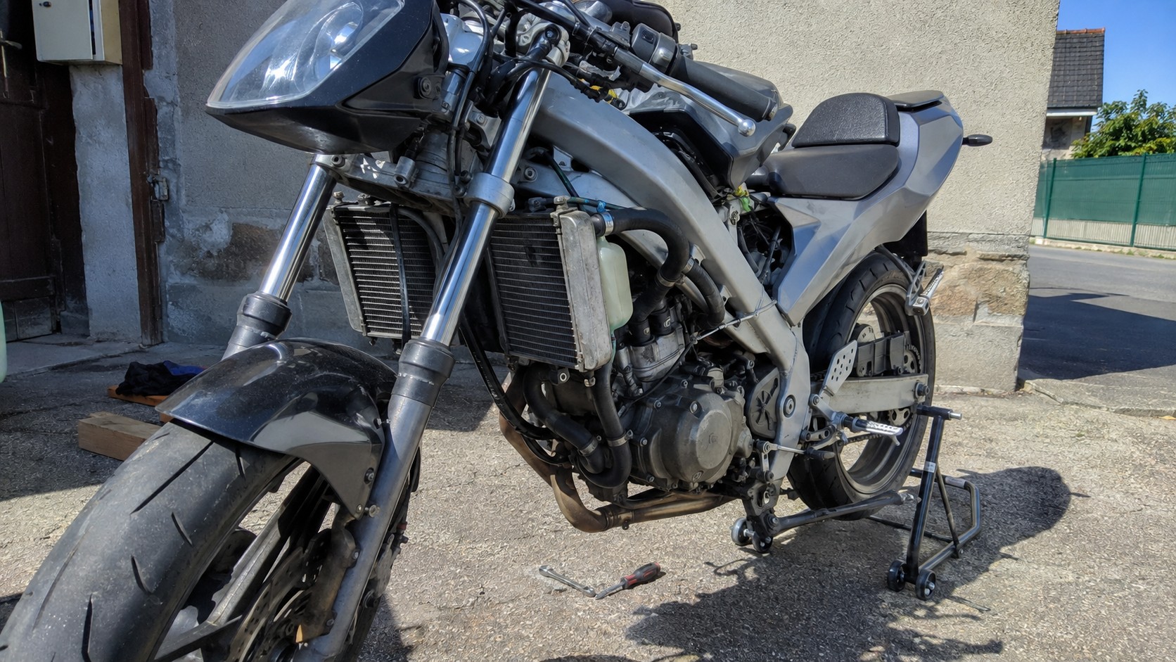 Moto grise partiellement démontée sur béquille d’atelier, vue trois-quarts avant gauche, avec deux radiateurs de refroidissement liquide exposés de part et d’autre de l’avant du moteur, durites, vase de liquide de refroidissement, fourche, roue avant et bloc moteur visibles, intervention mécanique dans une cour extérieure en plein soleil, illustration technique du principe du double radiateur pour moto et 50cc refroidi par liquide