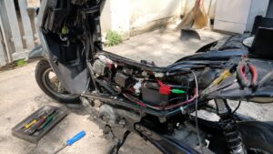 Scooter 50cc partiellement démonté en extérieur, selle et carénages retirés, faisceau électrique visible le long du cadre avec câbles rouges noirs verts, connecteurs blancs et verts, batterie 12 V avec borne positive protégée par capuchon rouge, boîtier électrique et régulateur à ailettes, câblage regroupé par colliers, moteur et transmission apparents, amortisseur arrière et roue visibles, pinces et tournevis posés sur le scooter et dans un bac métallique au sol, intervention de simplification et contrôle du faisceau électrique sous lumière naturelle ensoleillée