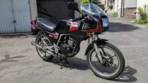 Kawasaki AR50 50cc vintage noire et rouge stationnée sur béquille centrale dans une rue résidentielle ensoleillée, vue trois quarts avant droit, carénage sportif avec bulle transparente, phare rectangulaire, clignotants orange, selle noire nervurée, moteur apparent, échappement latéral, jantes à bâtons et frein à disque avant, photographie documentaire réaliste au Canon EOS R6 en focale grand-angle 24–28 mm, lumière naturelle, netteté étendue, textures mécaniques détaillées, août 2026