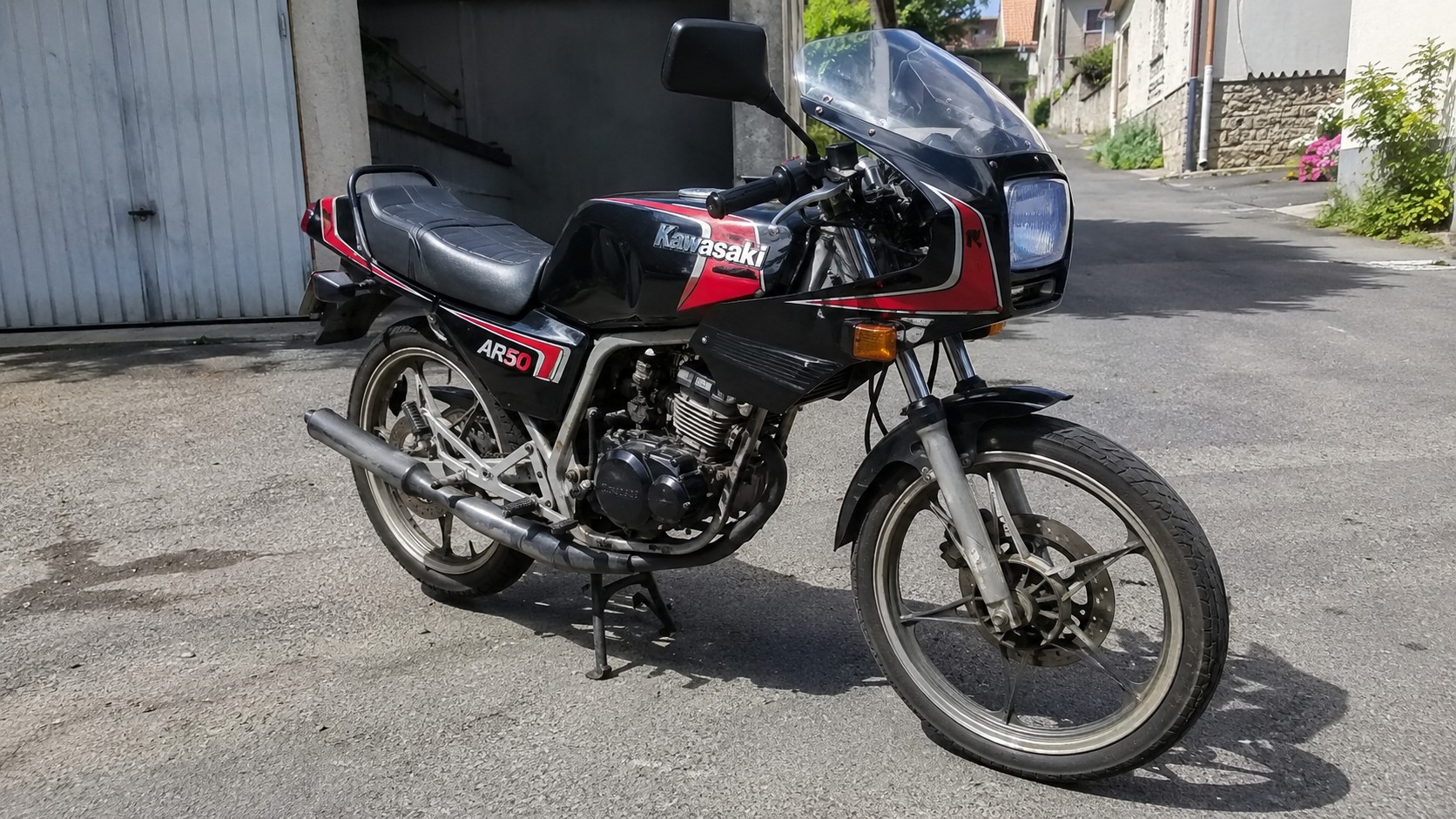 Kawasaki AR50 50cc vintage noire et rouge stationnée sur béquille centrale dans une rue résidentielle ensoleillée, vue trois quarts avant droit, carénage sportif avec bulle transparente, phare rectangulaire, clignotants orange, selle noire nervurée, moteur apparent, échappement latéral, jantes à bâtons et frein à disque avant, photographie documentaire réaliste au Canon EOS R6 en focale grand-angle 24–28 mm, lumière naturelle, netteté étendue, textures mécaniques détaillées, août 2026