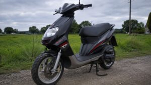 malaguti_50cc_scooter_noir_et_gris_accents_rouges_vue_trois_quarts_avant_gauche_stationne_sur_chemin_en_gravier_bequille_centrale_roue_avant_pneu_sculpté_jante_alliage_frein_a_disque_fourche_telesco­pique_optique_avant_guidon_selle_noire_marchepied_moteur_transmission_arriere_amortisseur_prairie_campagne_francaise_arbres_poteau_et_lignes_electriques_ciel_couvert_lumiere_naturelle_diffusee_photo_panasonic_gh4_24_28mm_2026_16_9