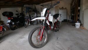 Moto 50cc type enduro blanche et rouge, vue trois-quarts avant, dans un garage avec un scooter et des vélos en arrière-plan