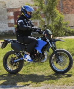 Mathéo Schweigart au guidon d’une moto 50cc supermotard yamaha bleue et noire, casque intégral bleu noir jaune, blouson et gants moto noirs, roues à rayons avec frein à disque avant, échappement 2 temps à détente visible, conduite sur terrain herbeux en extérieur par temps ensoleillé, 2026