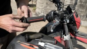 montage d’une poignée moto 50cc noire et rouge sur guidon côté accélérateur, mains installant le grip sur tube de gaz, levier de frein et commodo visibles, ancienne poignée usée, cutter, tournevis et flacon de montage posés sur la selle, moto rouge et noire en extérieur, lumière naturelle ensoleillée, prise de vue rapprochée Canon EOS R6, rendu documentaire réaliste, 2026