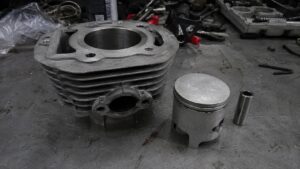 cylindre_50cc_moteur_2_temps_en_aluminium_à_ailettes_alésage_visible_piston_avec_segments_axe_de_piston_sur_établi_de_garage_mécanique_pièces_moteur_50cc_démontées_outils_et_traces_d_huile_panasonic_GH4_photo_réaliste_août_2026
