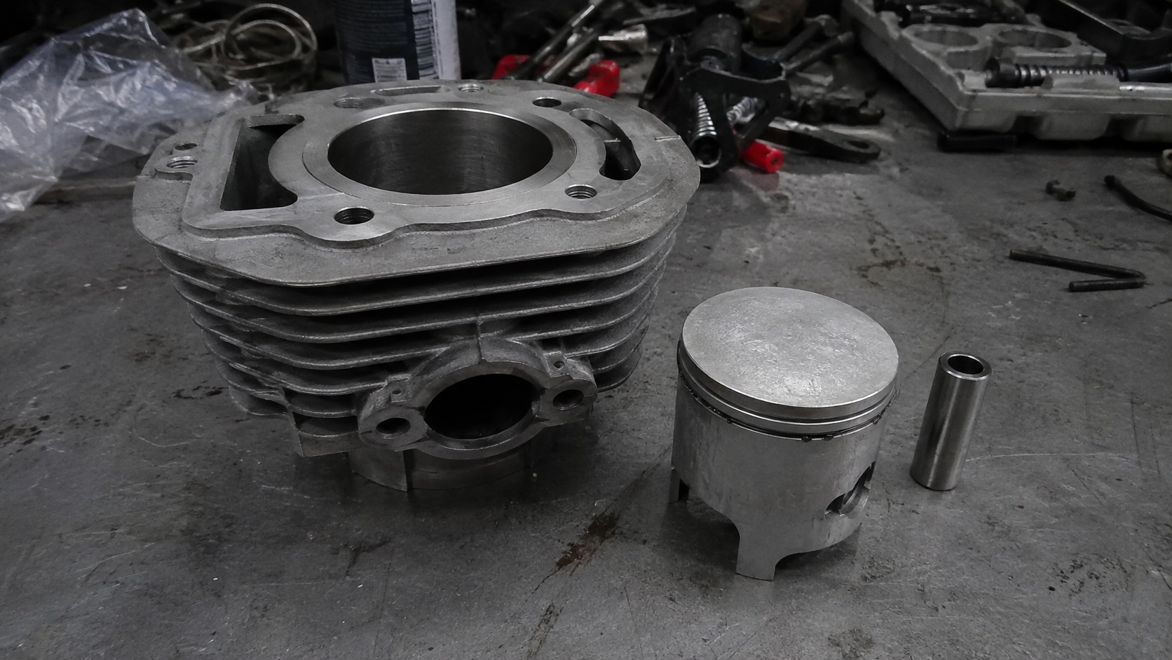 cylindre_50cc_moteur_2_temps_en_aluminium_à_ailettes_alésage_visible_piston_avec_segments_axe_de_piston_sur_établi_de_garage_mécanique_pièces_moteur_50cc_démontées_outils_et_traces_d_huile_panasonic_GH4_photo_réaliste_août_2026