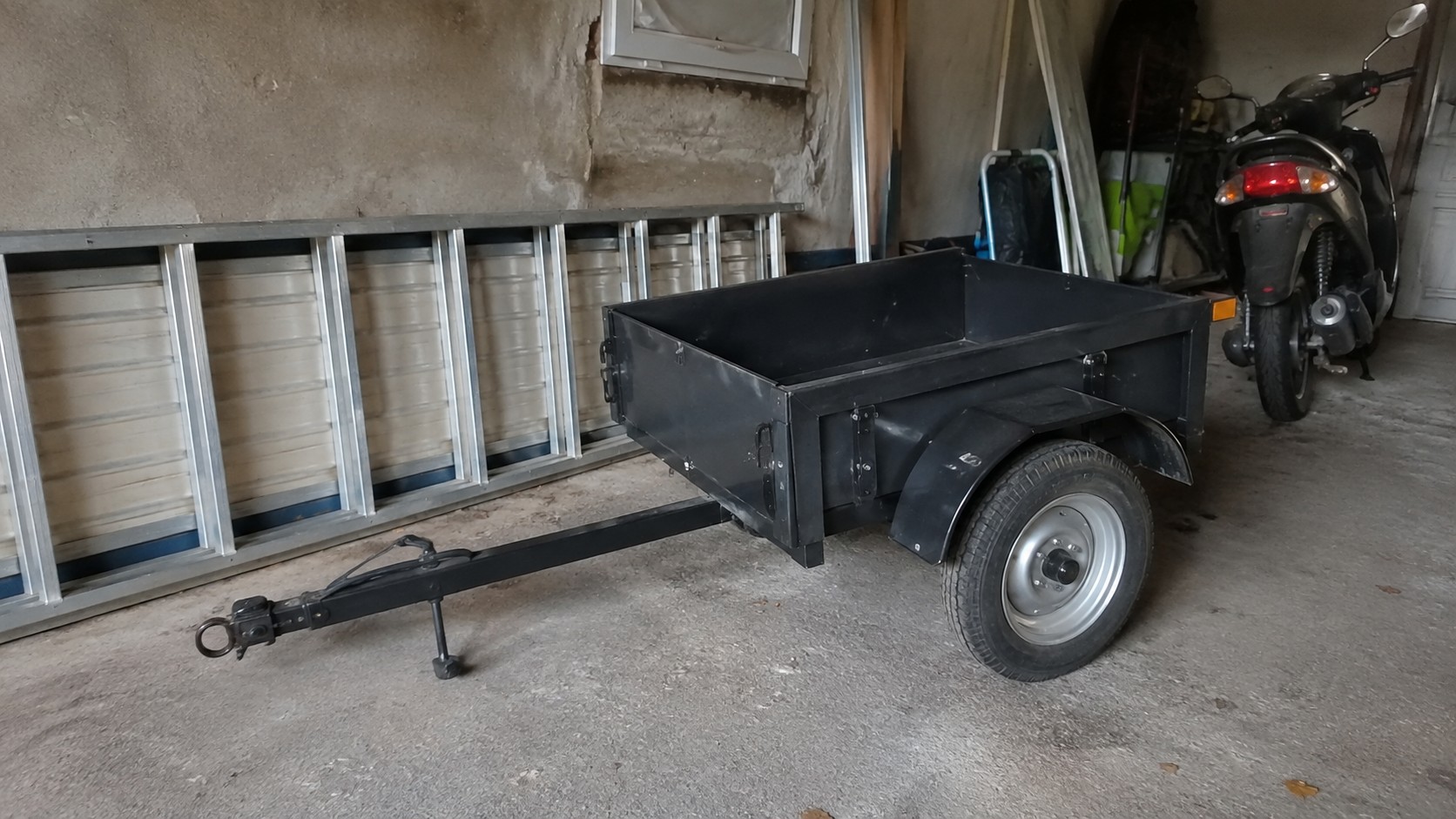 remorque_utilitaire_pour_scooter_50cc_noire_vue_latérale_dans_garage_français_châssis_métallique_benne_de_chargement_simple_essieu_roue_pneu_jante_garde_boue_flèche_d_attelage_béquille_de_support_scooter_garé_arrière_plan_2026