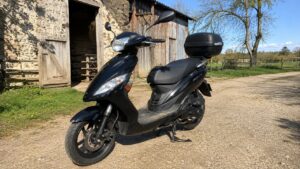 scooter_50cc_4_temps_noir_vue_3-4_avant_gauche_top_case_noir_retroviseurs_double_optiques_avant_roue_avant_frein_a_disque_fourche_avant_carter_moteur_4_temps_bequille_centrale_grange_agricole_en_pierre_portes_en_bois_cloture_champ_herbe_arbres_chemin_en_gravier_lumiere_naturelle_soleil_ombres_naturelles_ciel_bleu_texture_reelle_Panasonic_GH4_16-08-2026_16-9