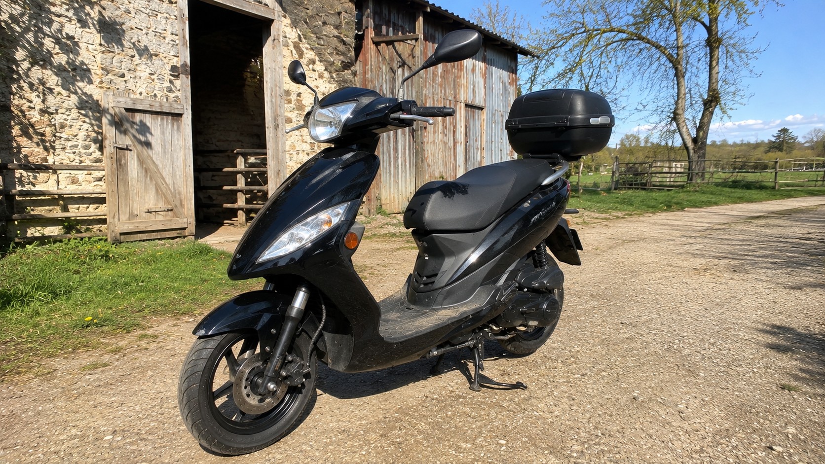 scooter_50cc_4_temps_noir_vue_3-4_avant_gauche_top_case_noir_retroviseurs_double_optiques_avant_roue_avant_frein_a_disque_fourche_avant_carter_moteur_4_temps_bequille_centrale_grange_agricole_en_pierre_portes_en_bois_cloture_champ_herbe_arbres_chemin_en_gravier_lumiere_naturelle_soleil_ombres_naturelles_ciel_bleu_texture_reelle_Panasonic_GH4_16-08-2026_16-9